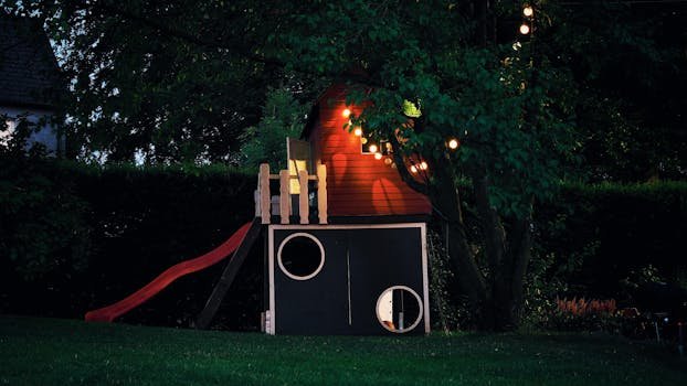 A whimsical treehouse illuminated by string lights in a lush backyard setting at dusk.